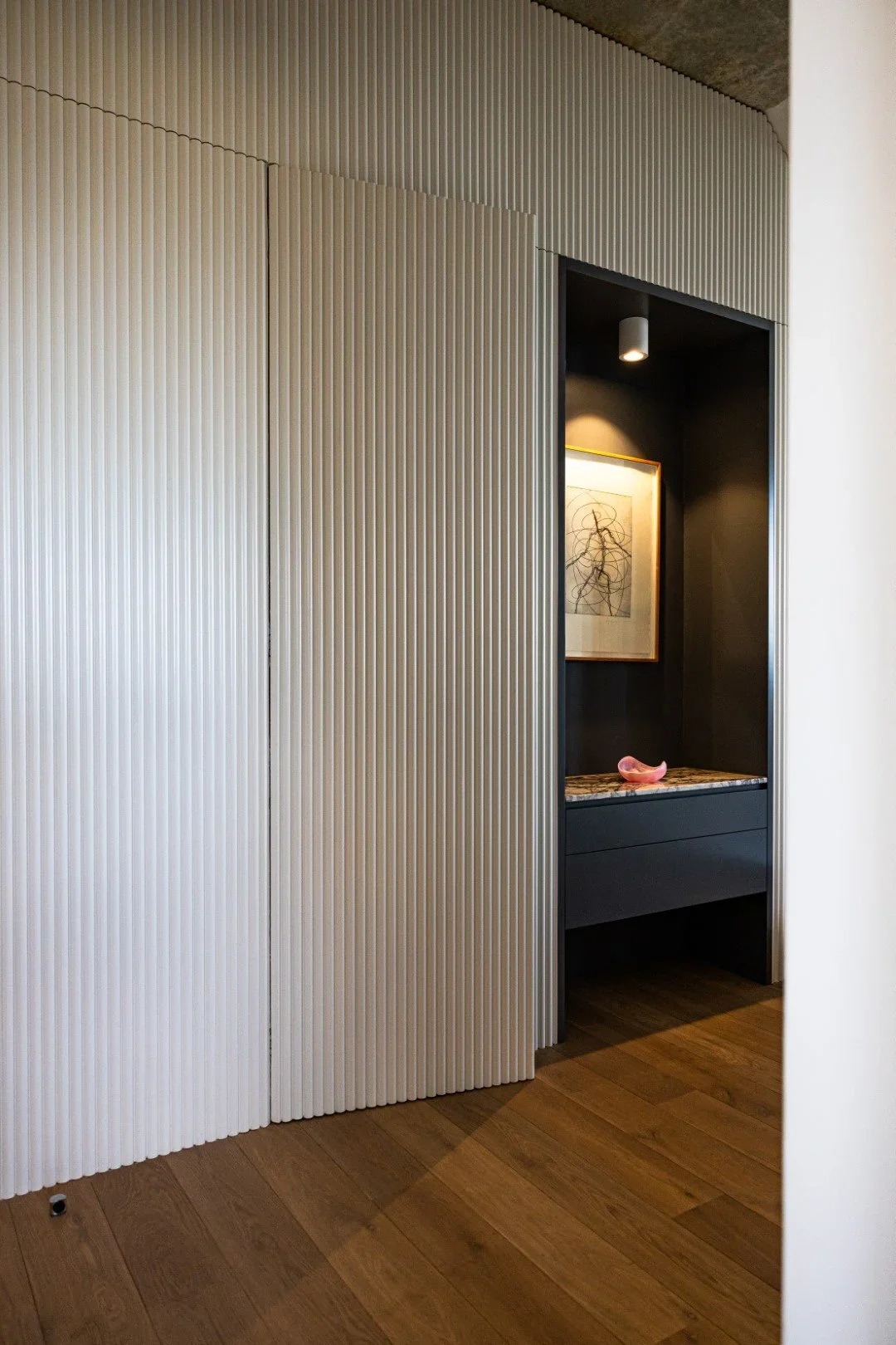 Interior corner with white textured sliding door, dark alcove with artwork and pink sculpture, wooden floor.