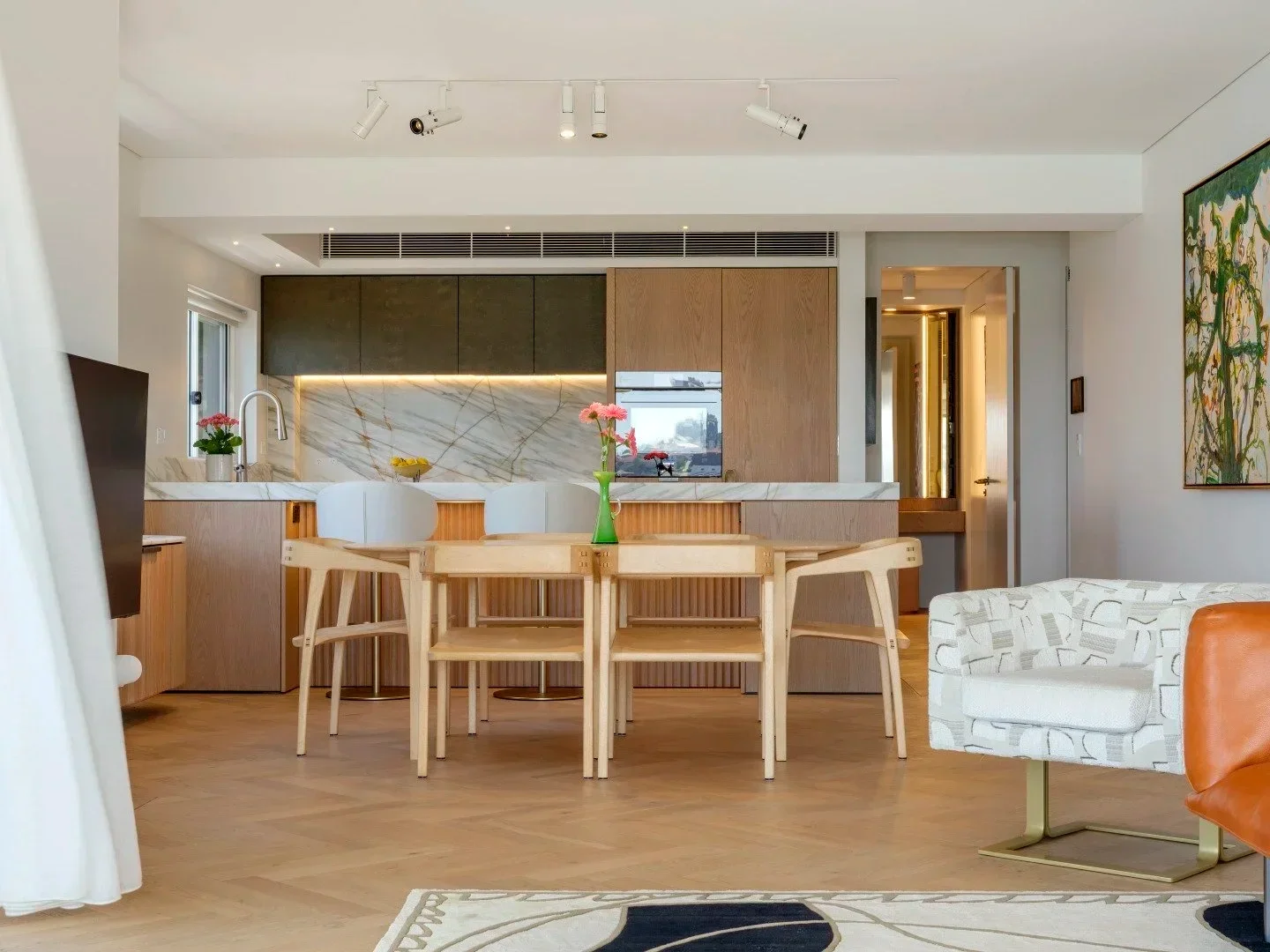 Modern living space with an open kitchen, dining area with a light wood table and chairs, a patterned white and beige armchair, and a patterned ottoman. The kitchen features a marble backsplash, wooden cabinetry, and track lighting.