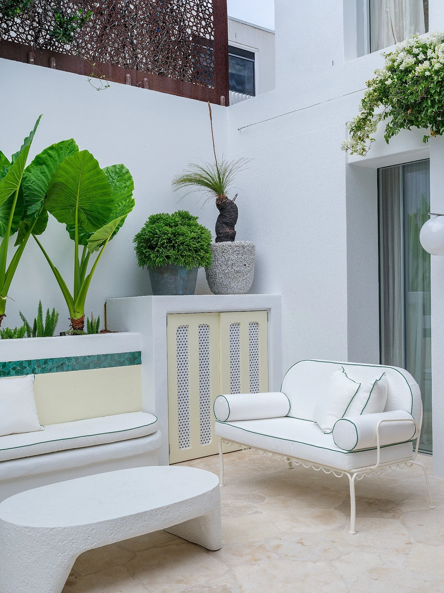 Outdoor patio with white and green furniture, lush plants, and trees illuminated by soft lighting at dusk.