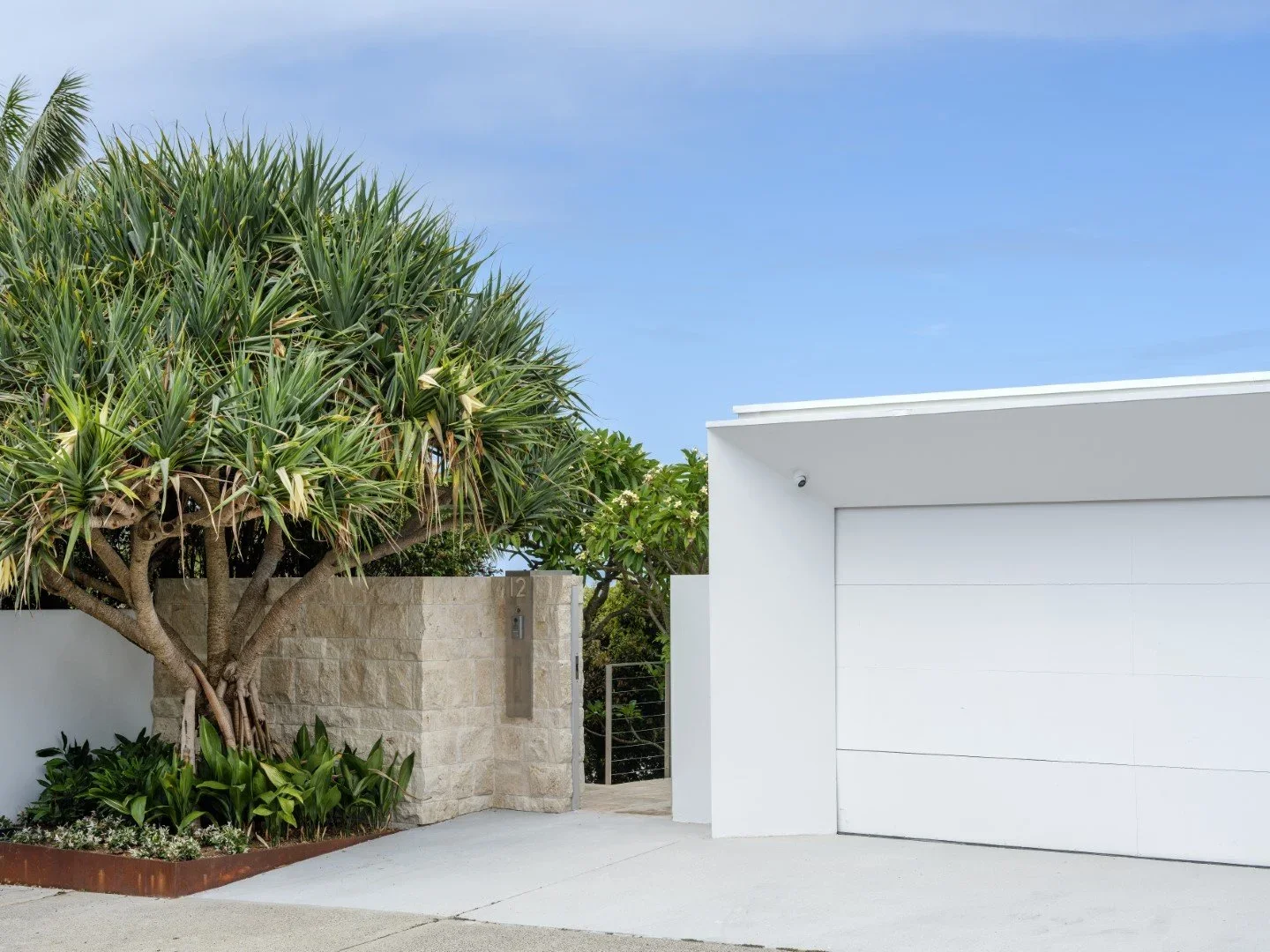 Modern house with white garage door, beige stone wall, and lush green plants including a large tree and smaller garden plants in front of the house, under a blue sky.