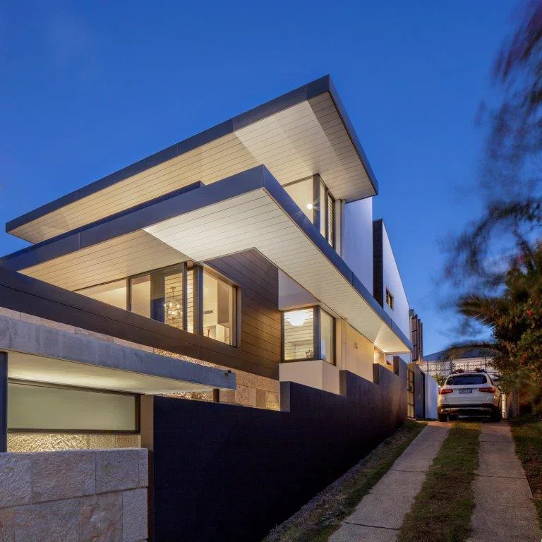 Modern multi-story house with large windows, geometric design, and exterior lighting at dusk, with a driveway and a white car parked outside.