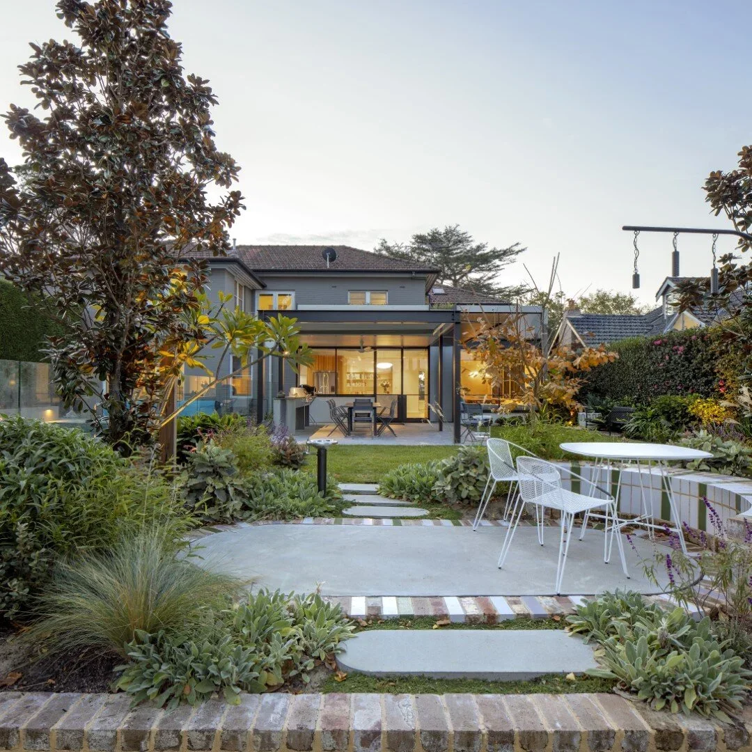 A modern backyard with a concrete patio, white outdoor furniture, lush green plants, and a well-lit house in the background during evening