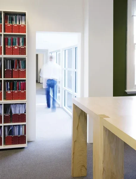 An office or library with a bookshelf filled with red and blue binders, a person walking in a bright hallway with large windows, and a light-colored reception or counter area.