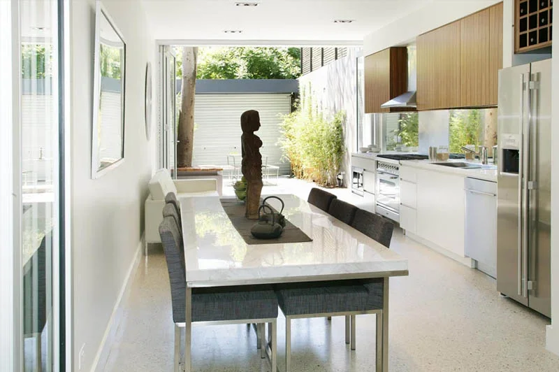 Modern kitchen and dining area with white cabinets, stainless steel appliances, wooden upper cabinets, a dining table with chairs, and a large window/door leading to an outdoor patio.