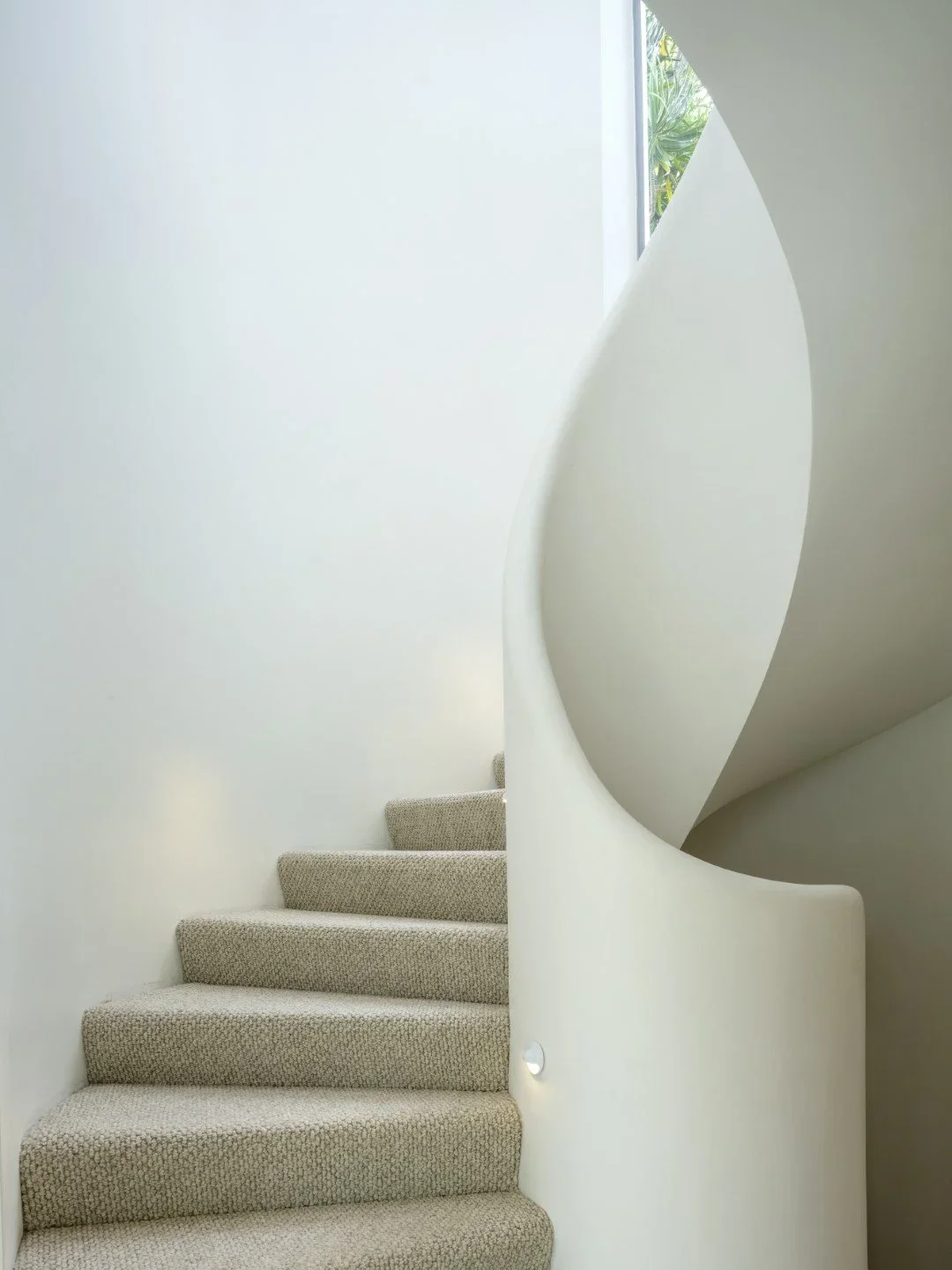 Modern interior staircase with beige carpeted steps and a white curved wall, illuminated by natural light from a window at the top.