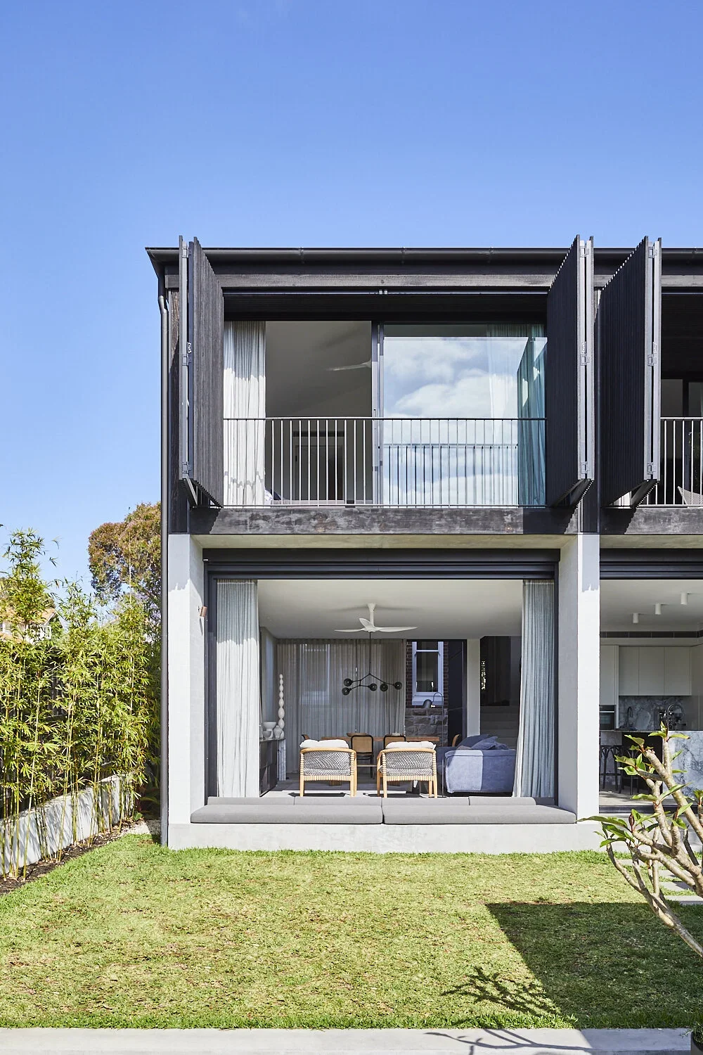 Modern two-story house with black and gray exterior, large sliding glass doors, open curtains revealing a dining area with chairs, a ceiling fan, and a living room with a couch inside, surrounded by a green lawn and bushes, under a clear blue sky.