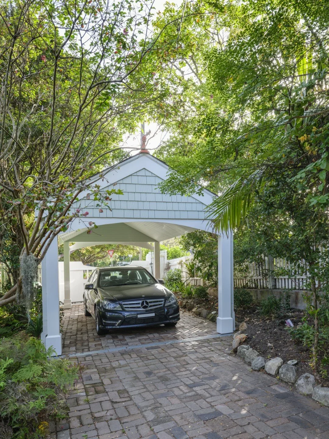 A black Mercedes-Benz parked under a white wooden carport, surrounded by lush green trees and plants, with a brick driveway and a white picket fence in the background.