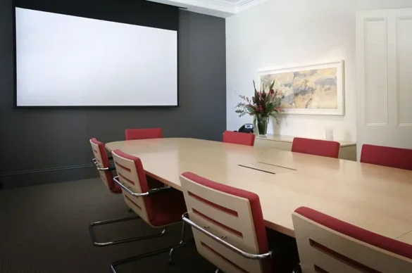 A conference room with a large wooden table surrounded by red and white office chairs, a whiteboard on the wall, and a vase of flowers on a side table.