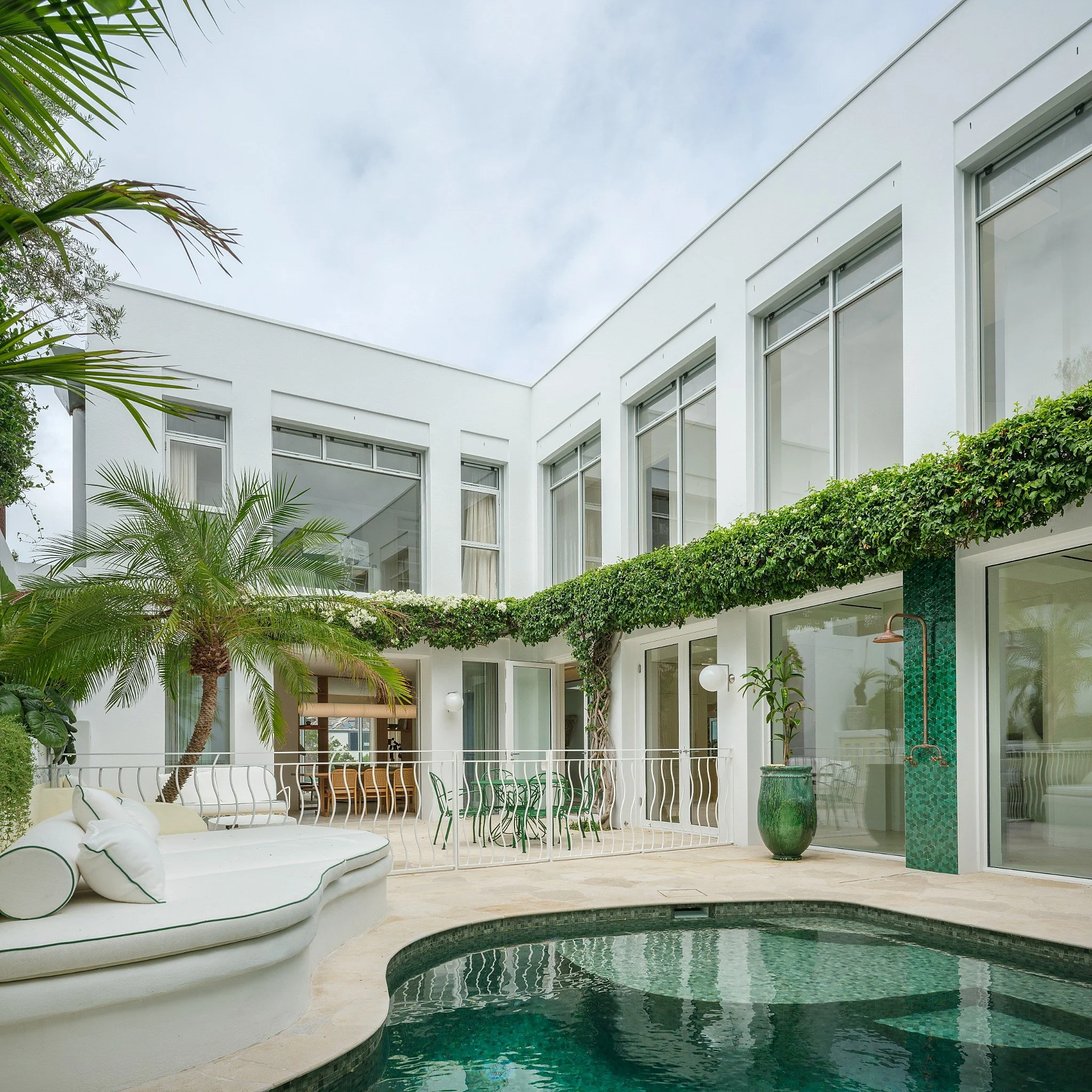 A modern white residential building with large windows, outdoor seating area, a swimming pool, lush green plants, and string lights, during dusk.