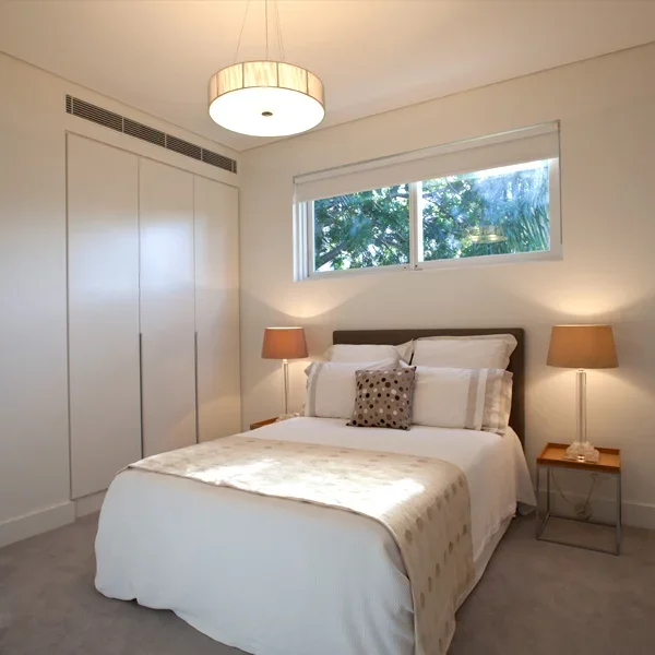 A bedroom with a white bed, two brown bedside lamps, a window with greenery outside, and a ceiling light fixture.