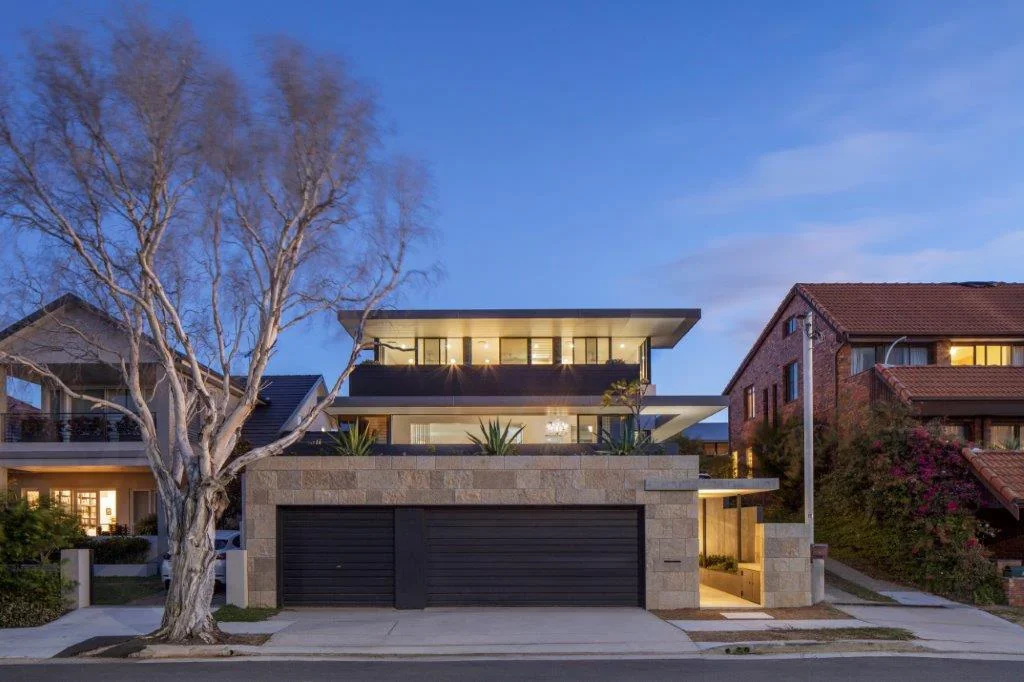 Modern multi-story house with large glass windows, stone facade, two garage doors, and a tree in front, during dusk.