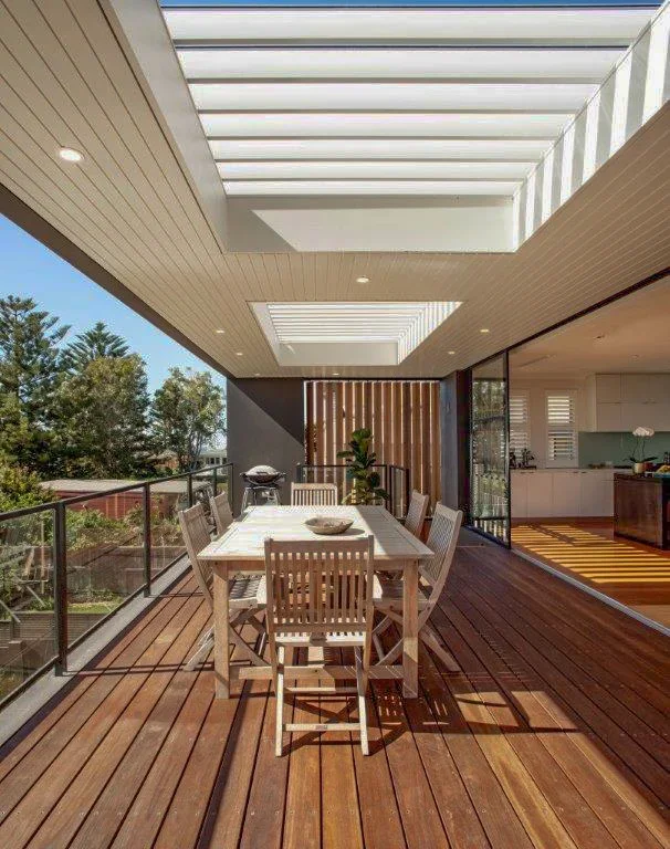 Outdoor balcony with a dining table and six chairs, glass railing, wooden flooring, and a modern ceiling with skylights, adjacent to a kitchen.