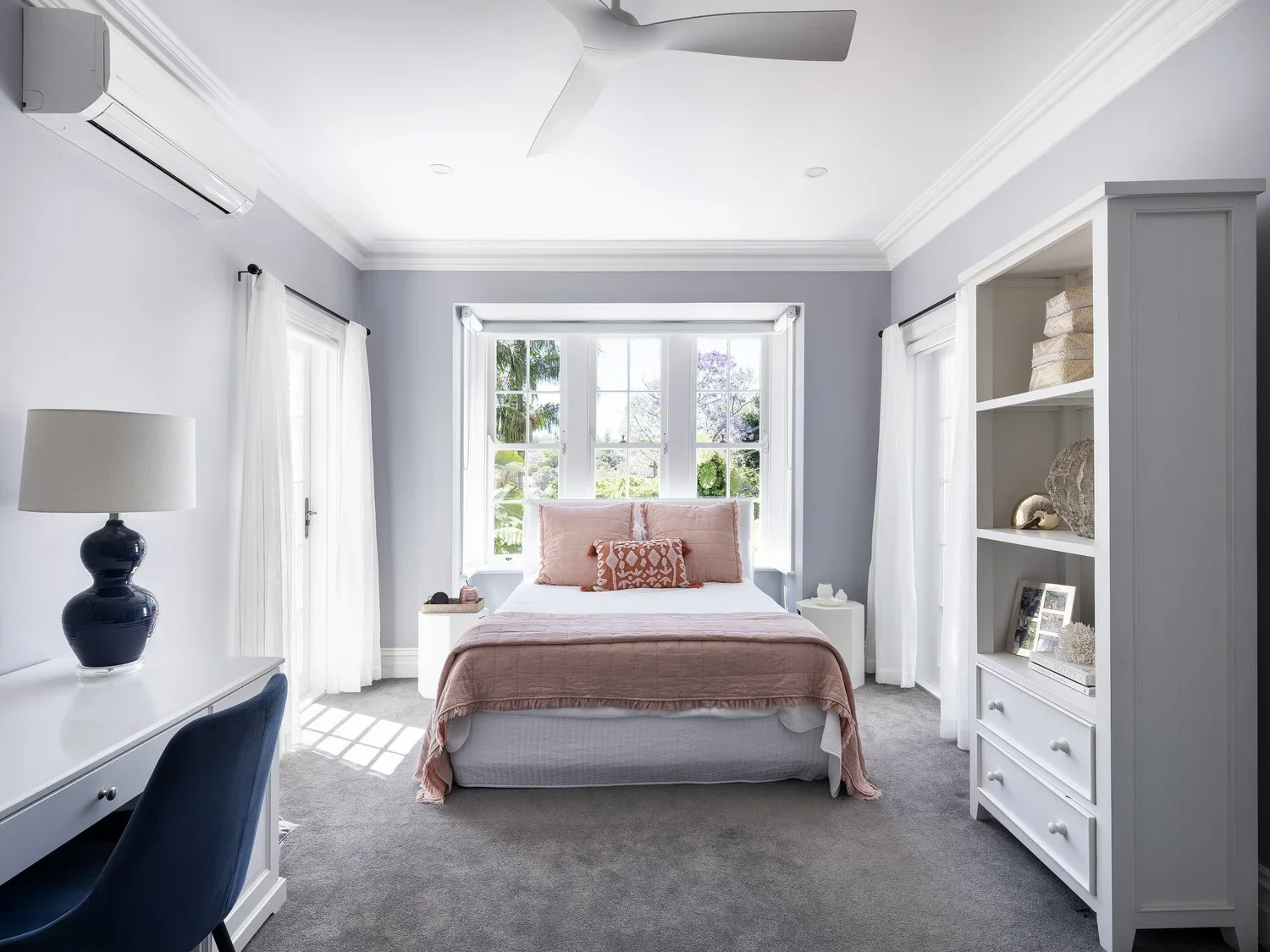 A bedroom with a bed in the center, pink bedding, white curtains, a window with greenery outside, a desk with a chair and a dark blue lamp, and a white bookshelf with decorative items.