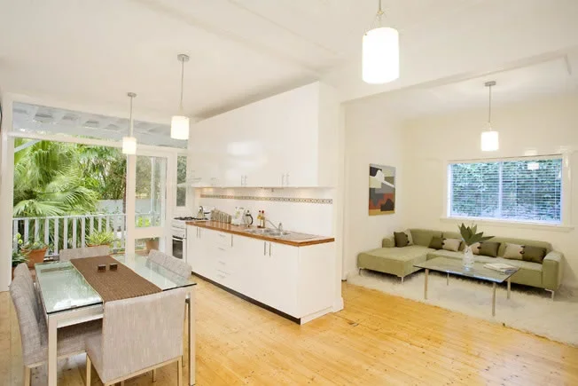 Open-concept living and dining area with a white kitchen, a glass dining table with six chairs, a green sectional sofa, large windows, and tropical outdoor scenery visible through a screened porch.