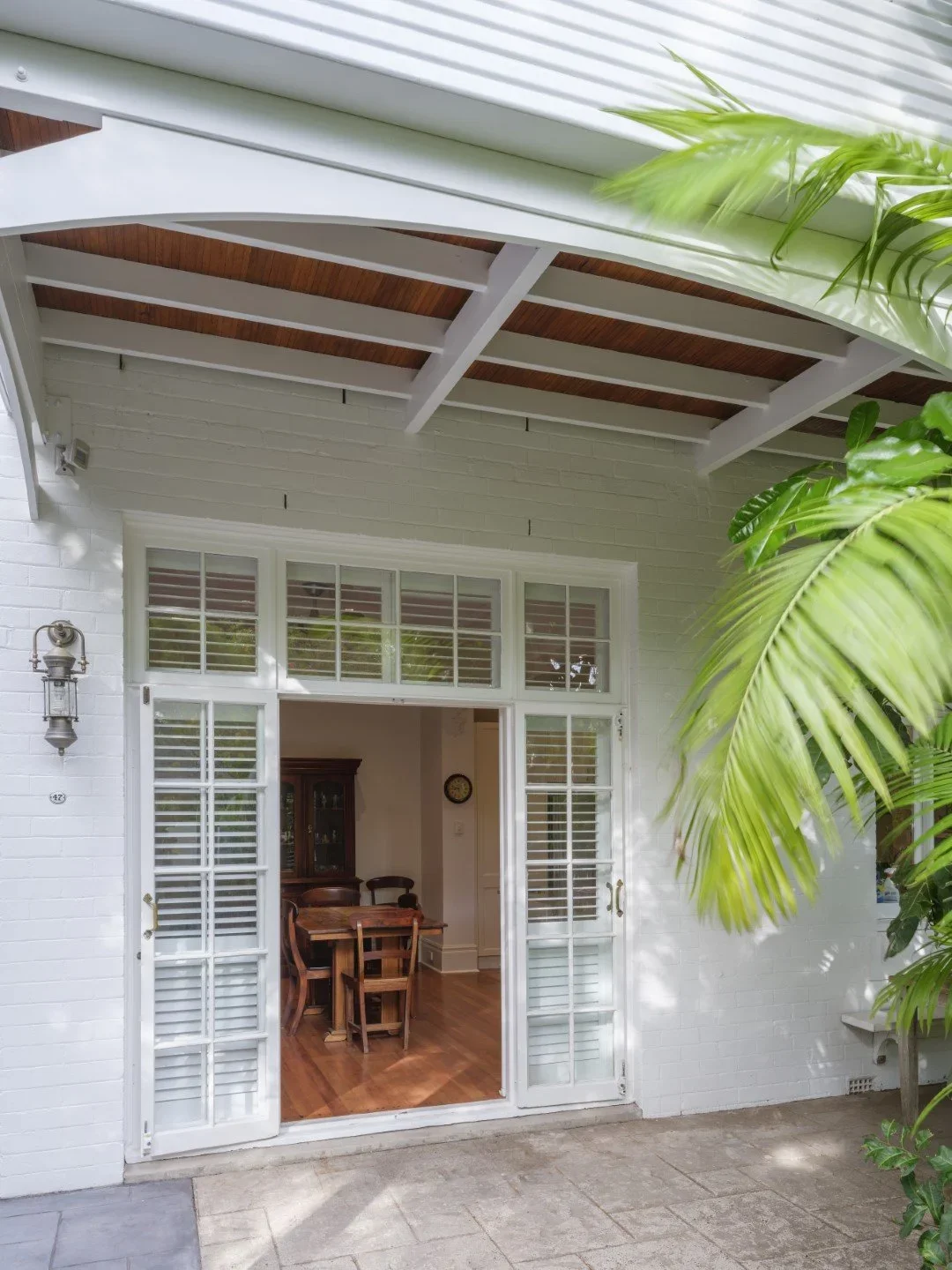 Open sliding glass door leading from a patio to a dining room with wooden furniture, white brick walls, and green plants outside.
