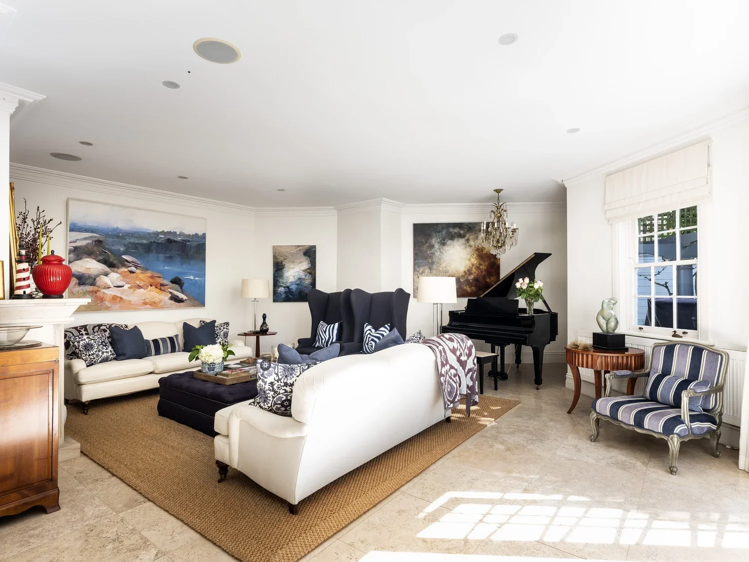 Spacious living room with white walls, beige tile floor, and large windows. Features a white sofa with black and white pillows, a black wingback armchair with striped pillows, a colorful area rug, and a grand black piano with a chandelier above. Deco