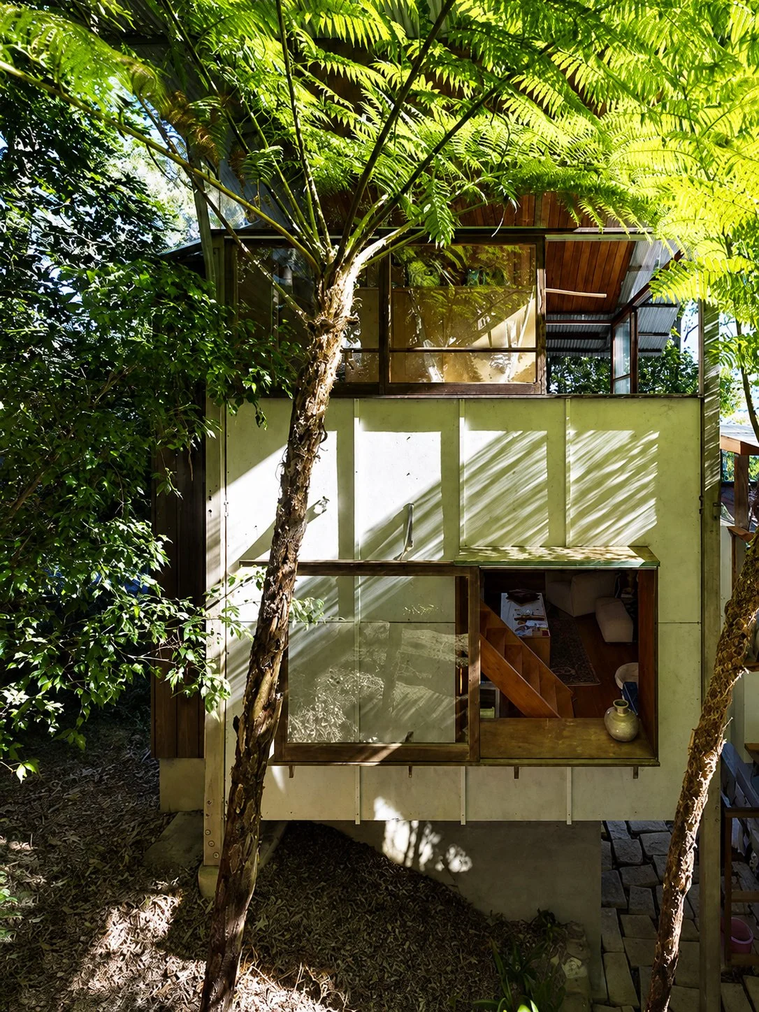 Designed by Peter Stutchbury. A modern two-story house with a curved roof surrounded by dense forest and ferns, built on a rocky hillside. The house has large glass windows and a wooden deck leading inside.