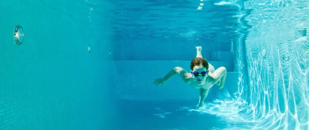 A young man swimming underwater in a swimming pool, wearing goggles.
