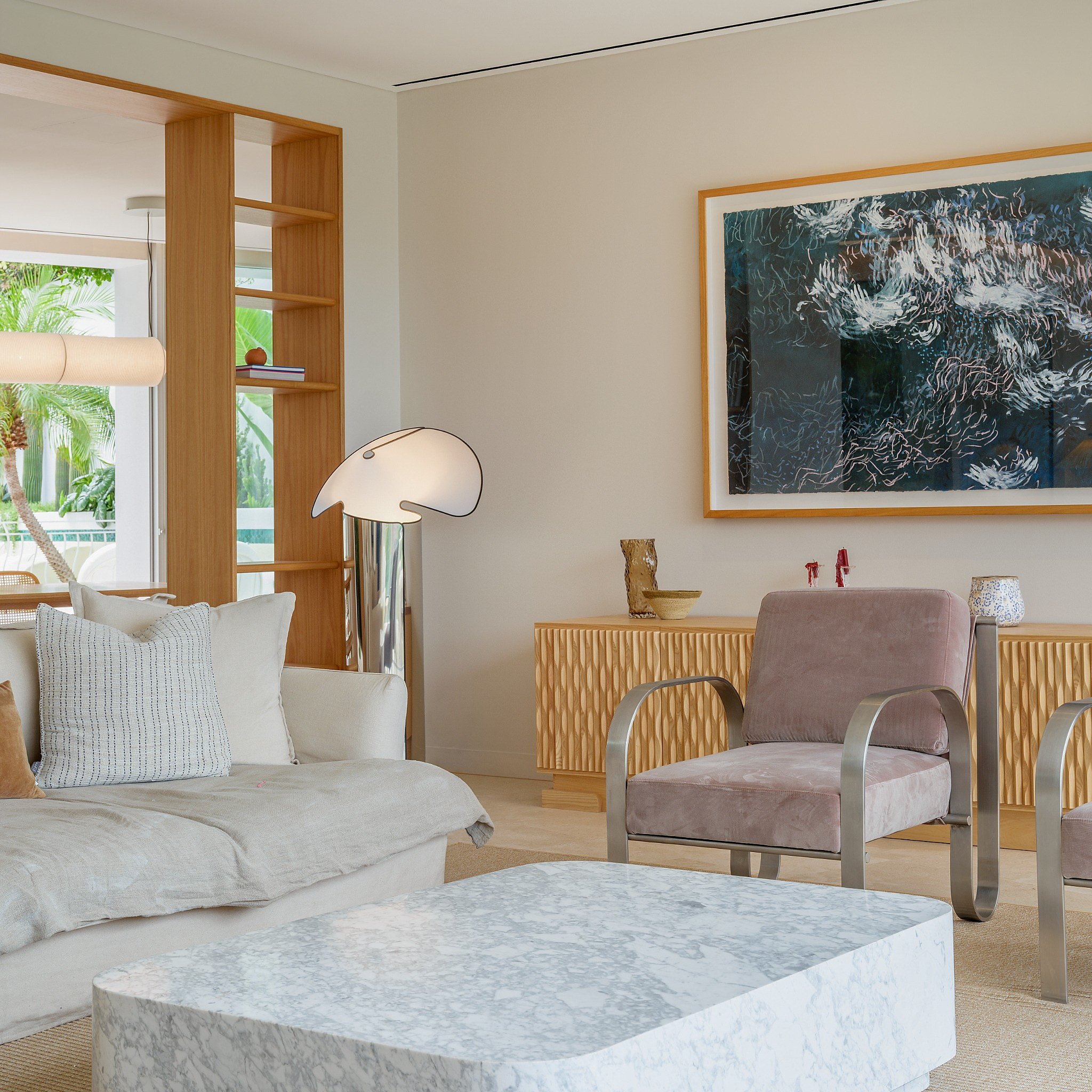 Living room with cream-colored sofa, marble coffee table, white armchair, fireplace, sliding glass door showing greenery, and minimalistic decor.