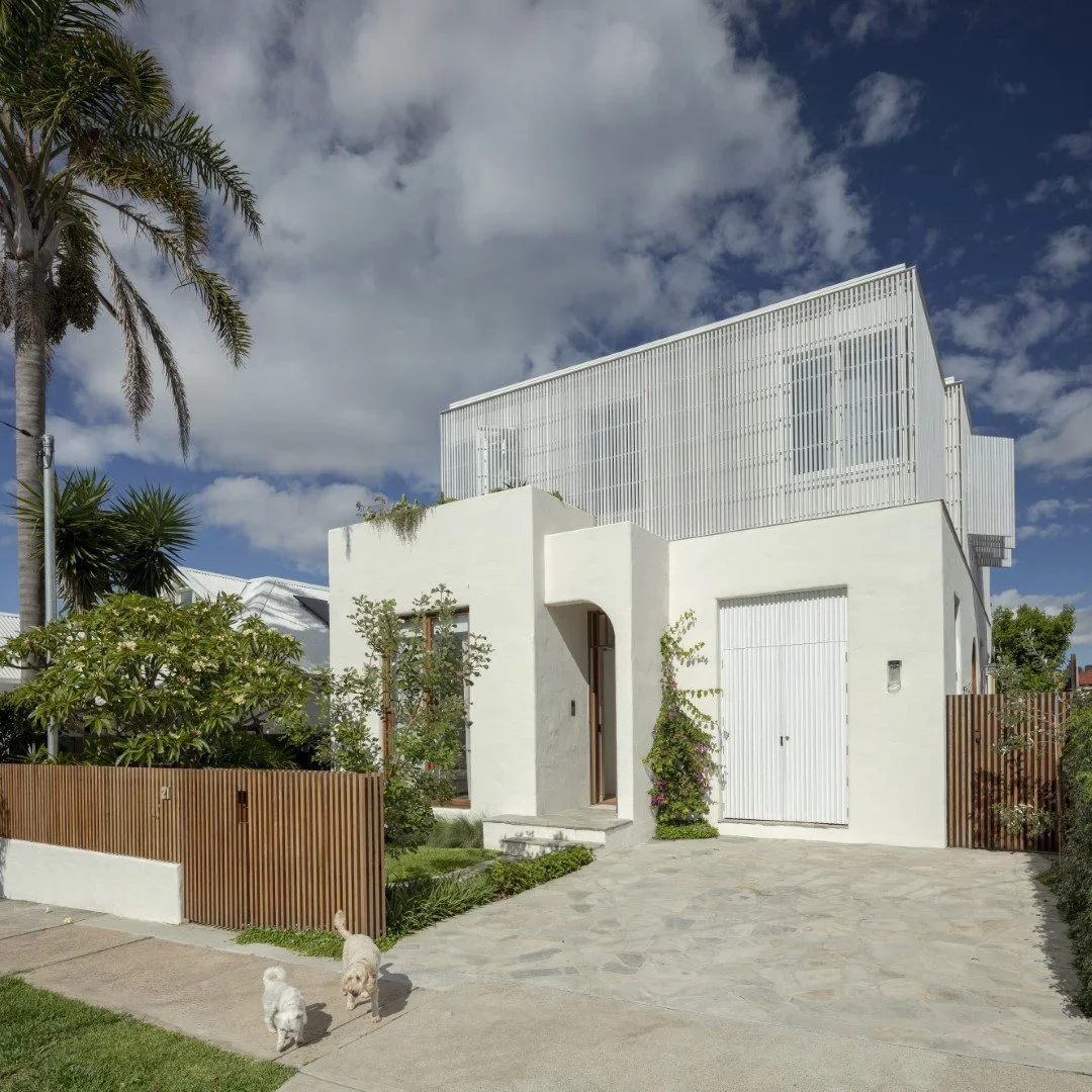 Modern white house with a wooden fence, surrounded by greenery, under a partly cloudy sky, with two dogs walking on the sidewalk in front.