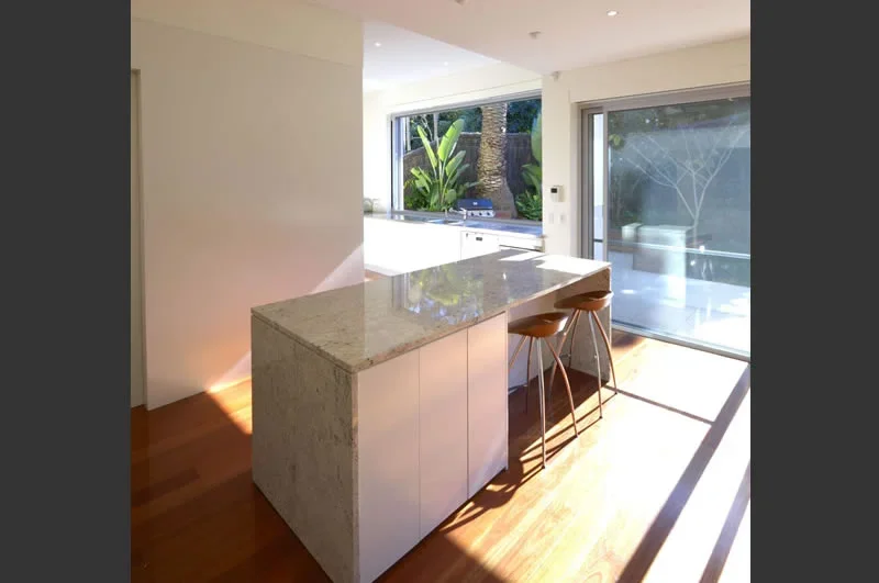 Modern kitchen with a marble island, two brown bar stools, large windows showing greenery outside, and sunlight streaming in.