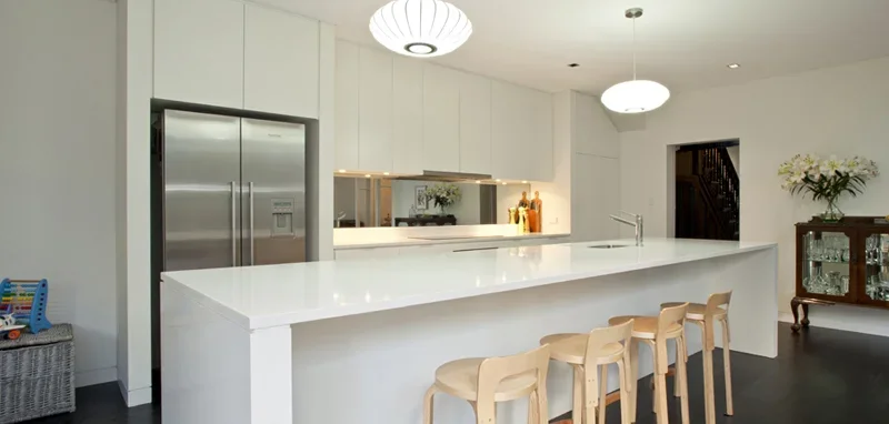 Modern kitchen with a large white island, beige bar stools, stainless steel refrigerator, white cabinets, and minimalist decor.