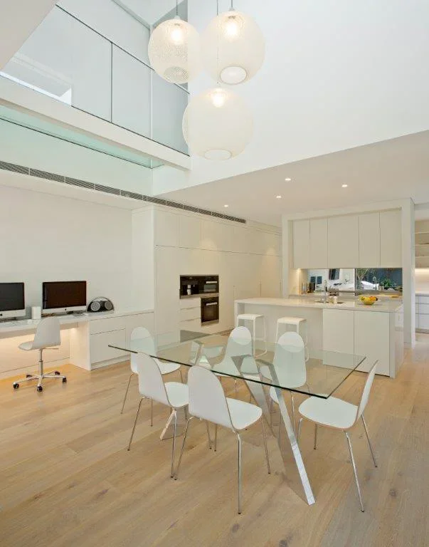 Modern, minimalist kitchen and dining area with white cabinets, bar stools, and a glass dining table with white chairs in a spacious, bright room.