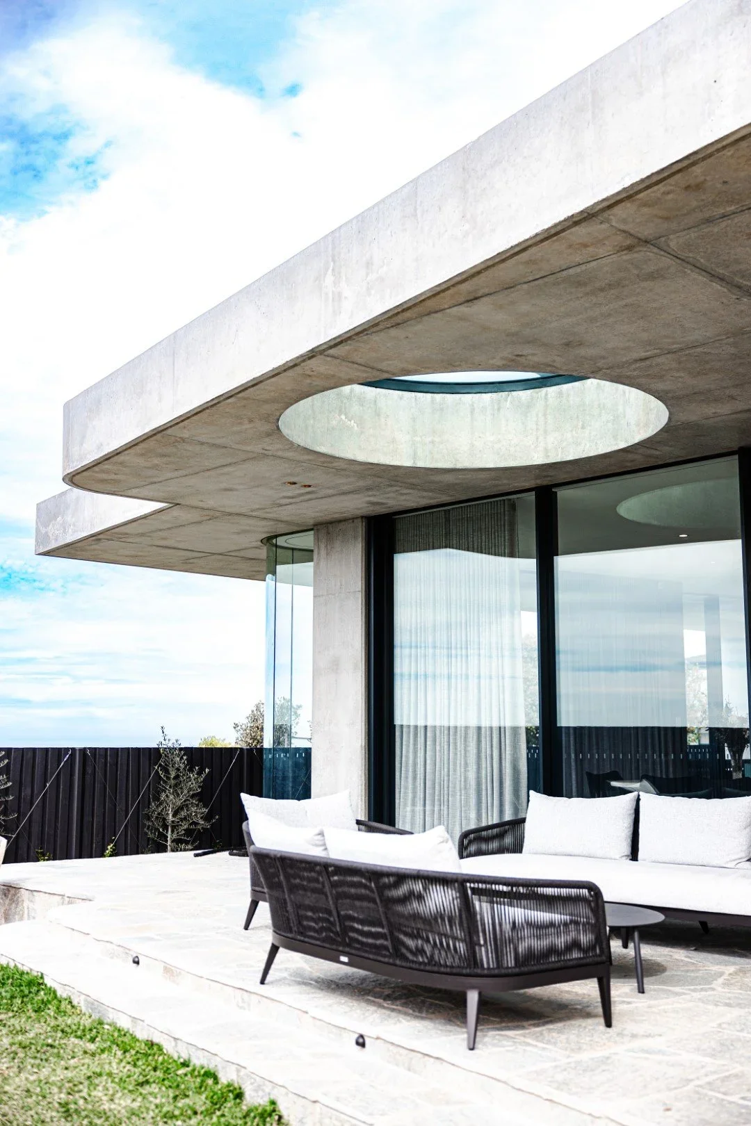 Modern house exterior with large glass doors, curved concrete roof with circular cutouts, and outdoor seating area with white cushioned furniture on a stone patio. Green grass and a wooden fence are visible in the background.
