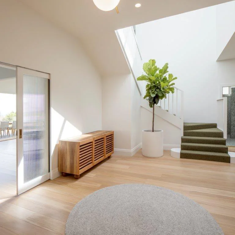 A bright, minimalist interior space with light wood flooring, a round gray rug, a wooden console, a large potted plant, and staircase with green-carpeted steps, illuminated by natural light from a skylight.