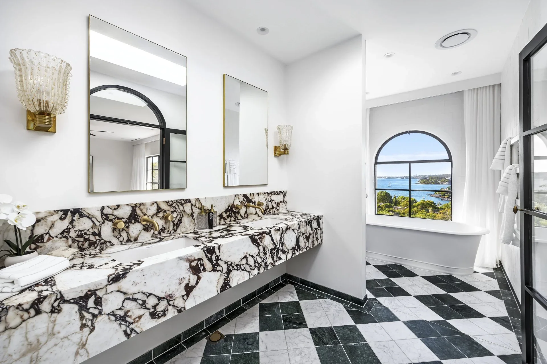 Modern bathroom with black and white checkered floor, a marble double vanity, mirrors, and a large window with a view of water and greenery.