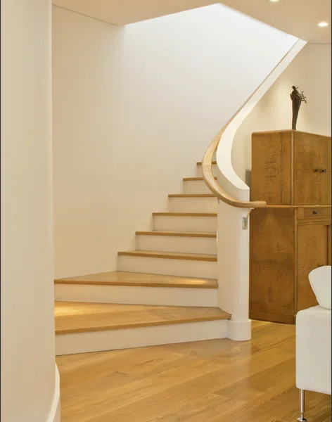 Interior view of a modern staircase with light wooden steps and a curved wooden handrail, next to a wooden cabinet with a decorative sculpture on top.