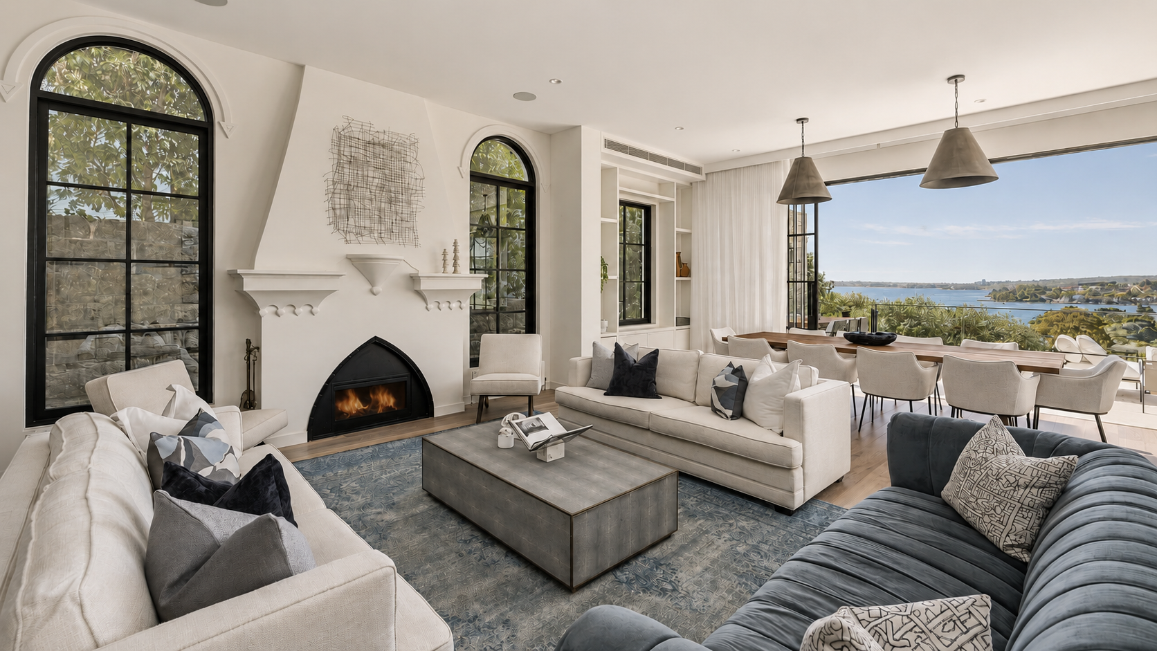 Living room with white sofas, a grey velvet sofa, a coffee table, and a fireplace with a fire. Large windows showing a view of water and trees outside, and a dining area with chairs and pendant lights.