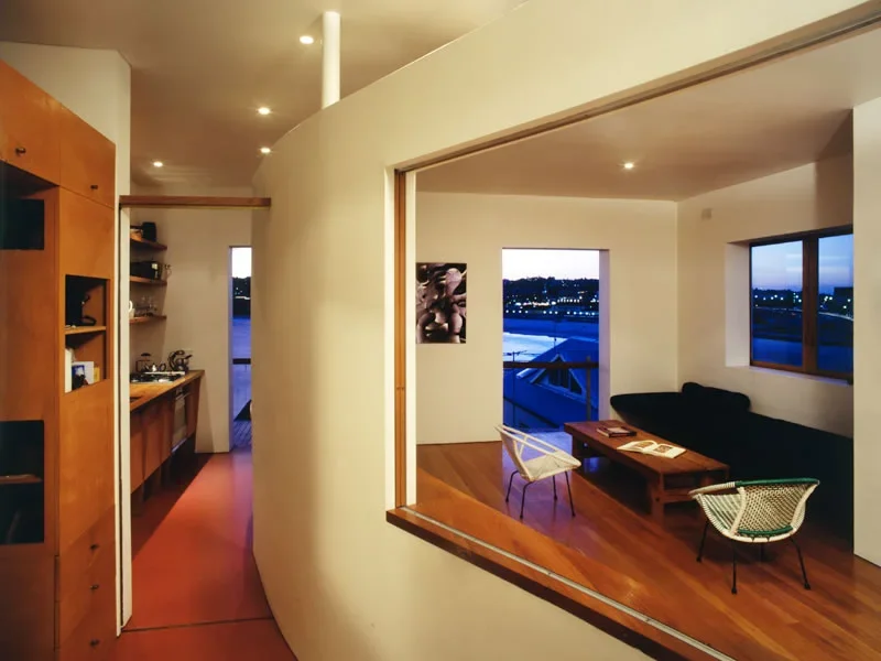 Interior view of a modern living room with wooden floors, large windows showing a city view at dusk, and a black sofa with a coffee table.