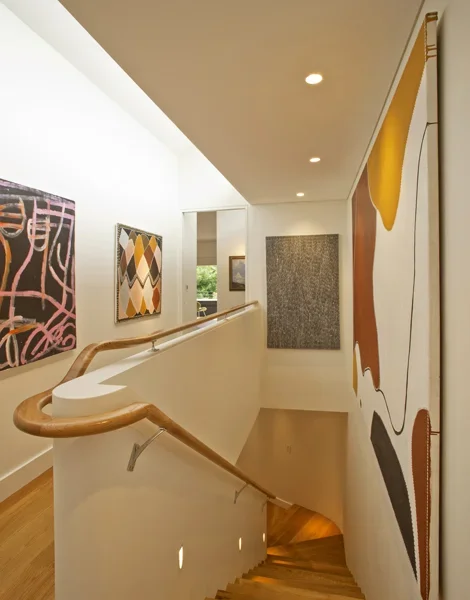 Art gallery hallway with various abstract paintings on white walls, wooden staircase with curved handrail, and a view into another room.