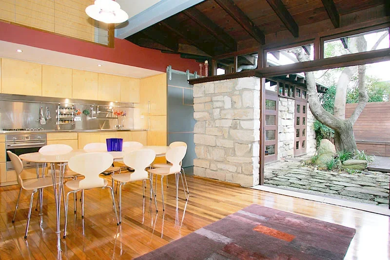 Open-concept kitchen and dining area with a wooden floor, modern white chairs around a round table, and a view of a stone patio and garden outside.