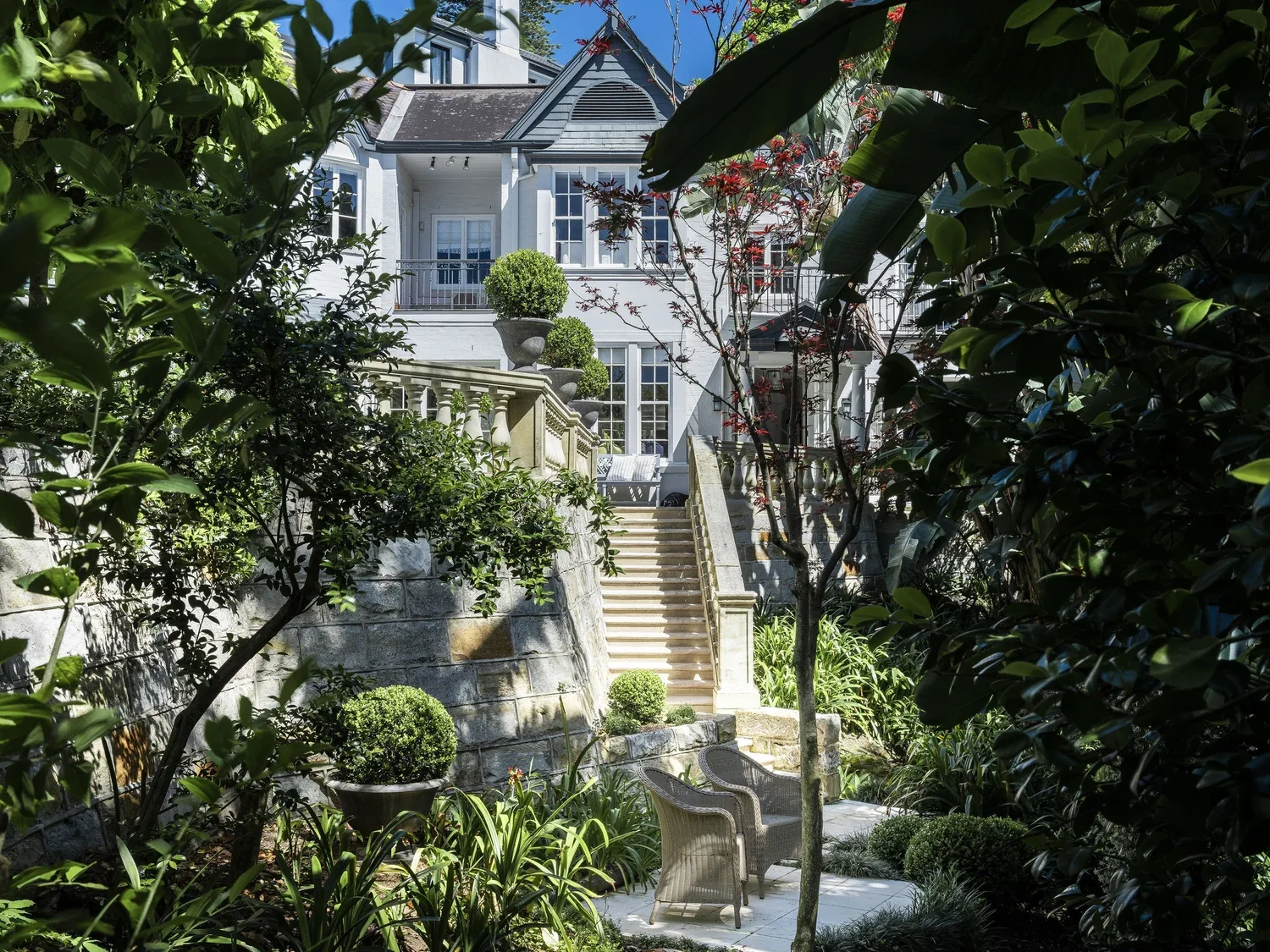 A lush garden leading up to a white house with steps, trees, shrubs, and outdoor seating.