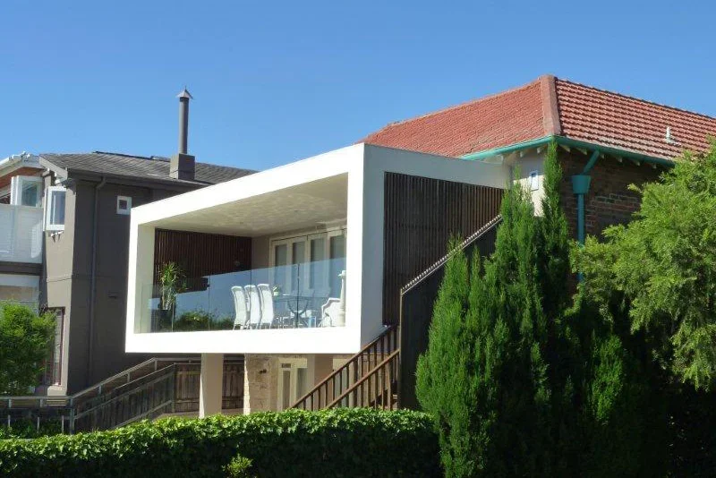 Modern house with a white rectangular balcony, wooden stairs, green trees, and a red tile roof under a clear blue sky.
