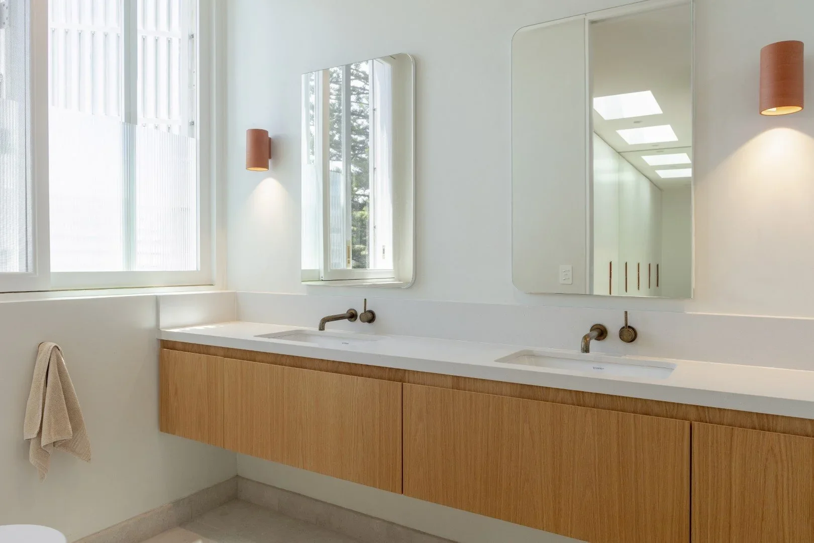 Modern bathroom with large windows, two mirrors, wall-mounted lights, and a wooden vanity with dual sinks.