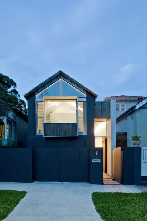 Front view of a modern black house with large windows, a small front yard, and a paved walkway at dusk.