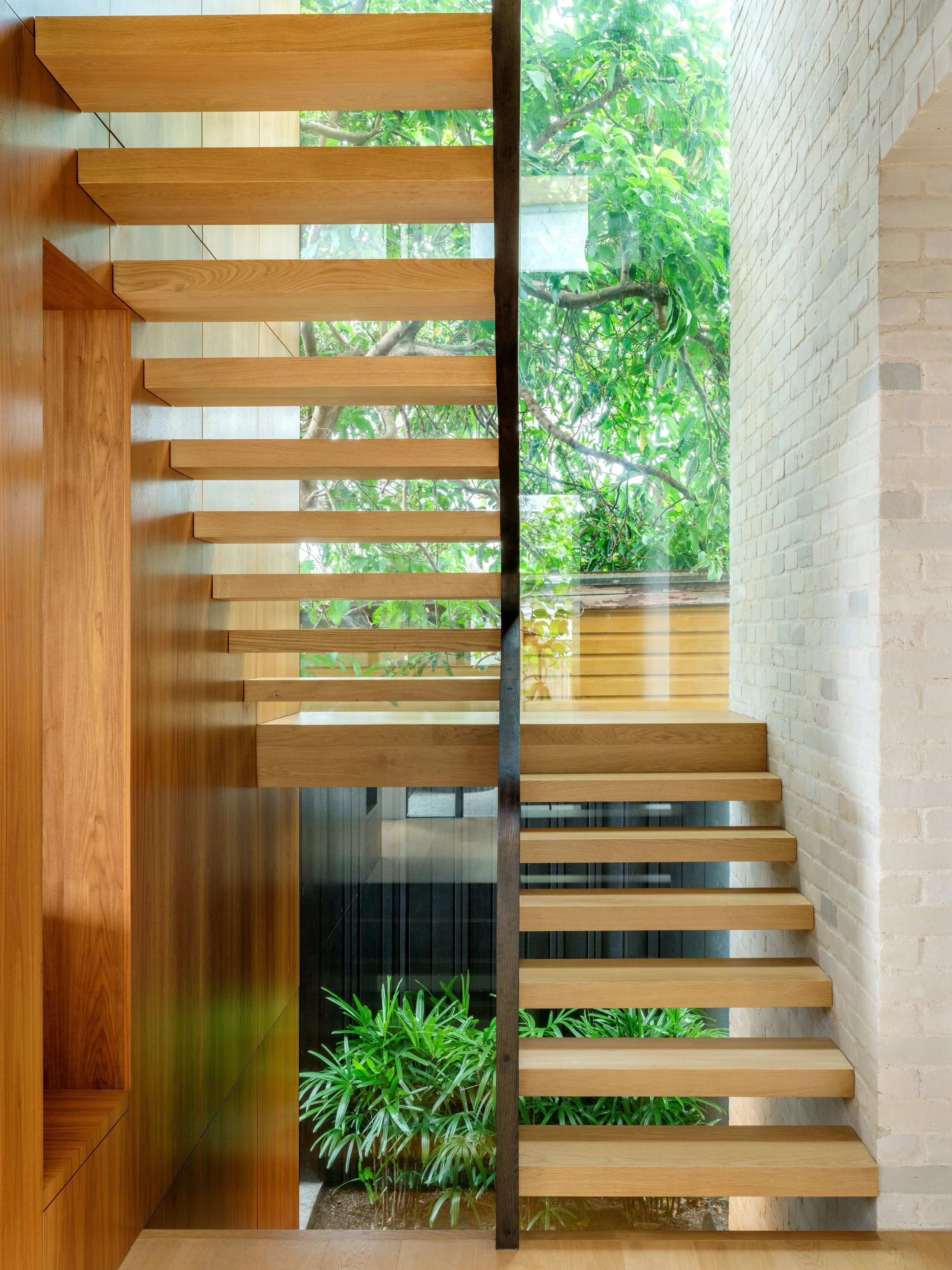 Open timber staircase at a Bronte home, with a dark steel balustrade, pale brickwork and a planted courtyard.