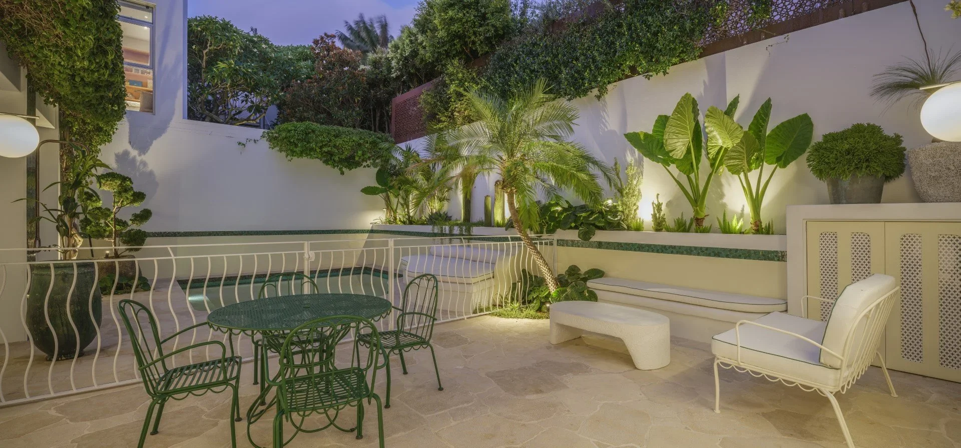 Outdoor patio with white and green furniture, lush plants, and trees illuminated by soft lighting at dusk.