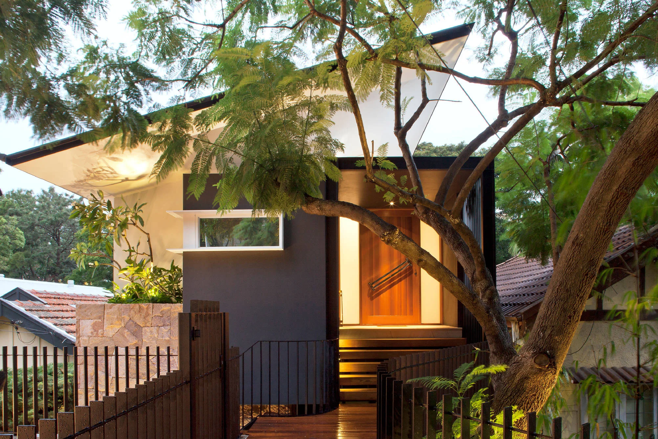 Modern house with a wooden front door, illuminated stairs, surrounded by lush green trees and a black iron fence.