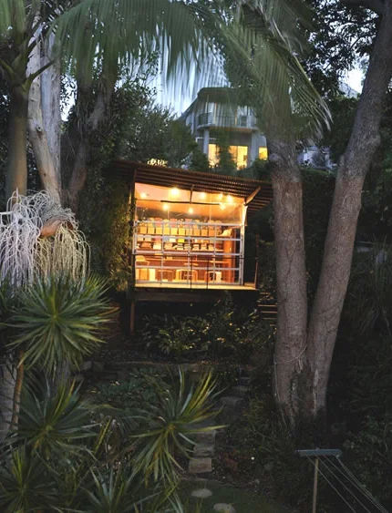 An outdoor modern structure with large glass windows, illuminated with warm lights, situated among trees and plants at dusk.