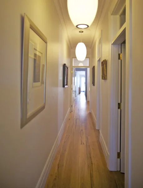 A narrow hallway with white walls, framed artwork, wooden flooring, and two hanging ceiling lights leading to a door at the end.