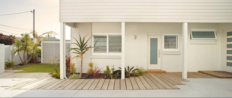 Modern white house with wooden deck and small landscaped garden with plants and shrubs.