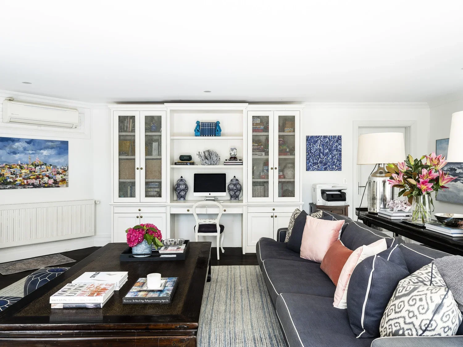 Living room with white built-in bookshelf, dark gray sofa with pink and patterned pillows, coffee table with books and flowers, artwork on walls, and a window air conditioning unit.