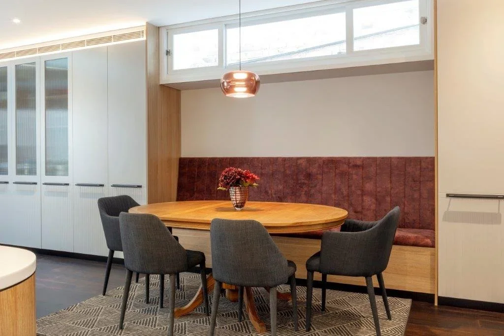 A dining area with a round wooden table, four gray upholstered chairs, a built-in bench with red upholstery, a vase with flowers, a window, and modern cabinetry.