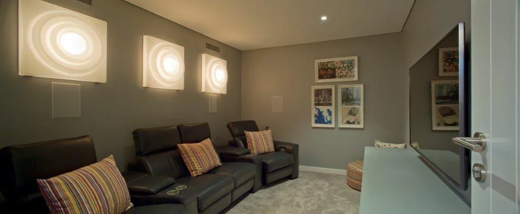Living room with three black leather chairs, each with a striped pillow, a flat-screen TV on a white stand, gray walls decorated with abstract art, and ambient wall lighting.