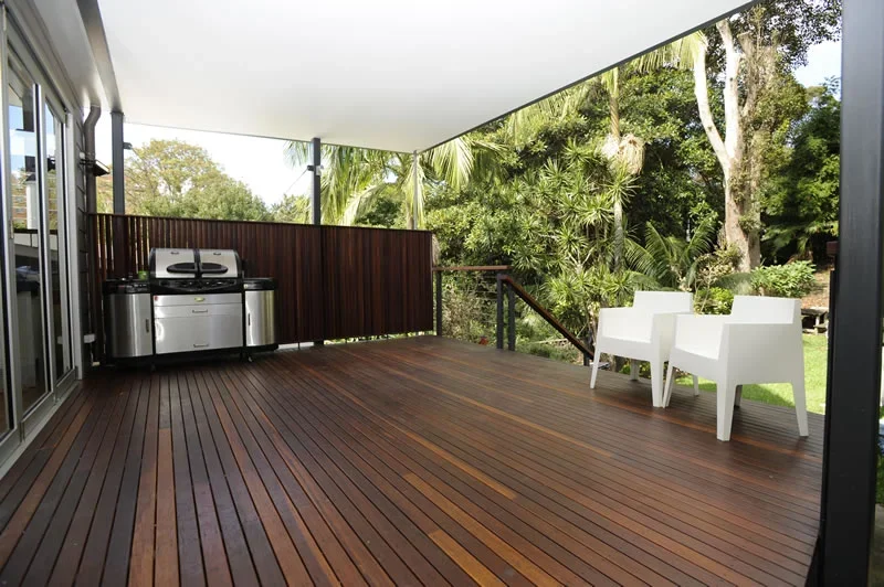 A spacious outdoor deck area with wooden flooring, two white chairs, and a grill, surrounded by lush green trees and plants.