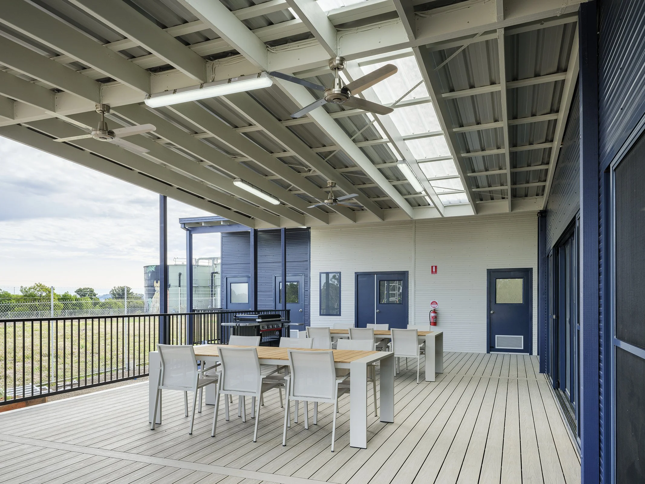 Empty outdoor balcony or patio with wooden flooring, white tables and chairs, enclosed with a metal railing, with a blue building wall, doors, and ceiling fans, under a translucent roof panel.
